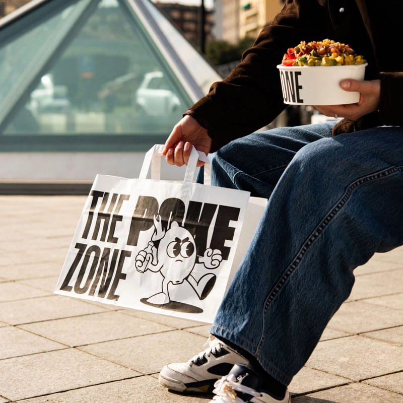 Person holding a custom-printed paper bag with 'The Poke Zone' logo, along with a branded bowl, showcasing takeaway packaging with logo.